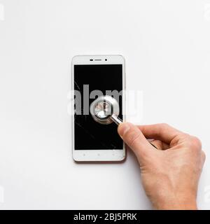 Nahaufnahme der Hand des Mannes, der Stethoskop auf gebrochenem Smartphone hält, Blick von oben, auf weißem Hintergrund. Kopierbereich. Smartphone mit cra Stockfoto