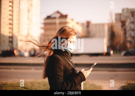 Junge Frau trägt schützende Gesicht medizinische Maske, während mit Smartphone zu Fuß auf der Straße in der Stadt Stockfoto
