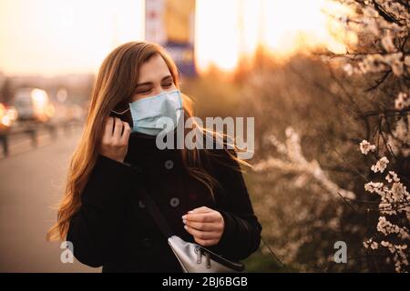 Glückliche junge Frau, die auf Gesicht medizinische Maske während des Stehens auf der Straße in der Stadt während Sonnenuntergang im Frühling setzt Stockfoto