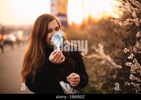 Glücklich fröhlich junge Frau Entfernen Gesicht medizinische Maske während auf der Straße in der Stadt während des Sonnenuntergangs im Frühjahr stehen Stockfoto