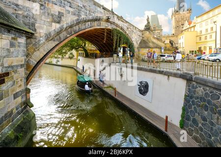 Ein Kanal mit einem Touristenboot unter einer Brücke auf der Kampa Insel in Prag, Tschechien mit dem Brückenturm der Kleinseite in der Ferne Stockfoto