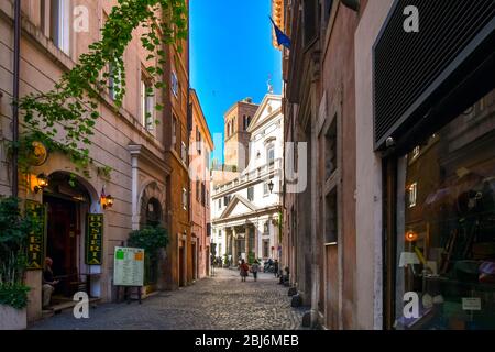 Zwei Frauen gehen eine schattige Gasse hinunter in Richtung der Kirche Sant' Eustachio mit dem Kopf eines weißen Hirsches, der sein Kreuz im historischen Rom hält. Stockfoto