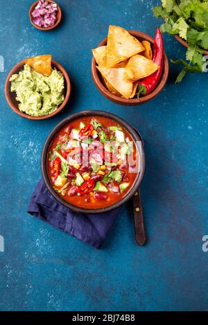 Mexikanische schwarze Bohnensuppe mit Tomaten, Avocado und Totopos. Blauer Hintergrund, Draufsicht. Stockfoto