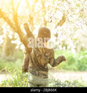 Back view Portrait von niedlichen schwarzen Haaren Jungen mit Spaß in der Mitte der Blüte Garten. Kind trifft weiße Blumen mit einem Stock. Im Freien, kopieren Raum, grün und weiß Frühlingsblumen Hintergrund. Stockfoto