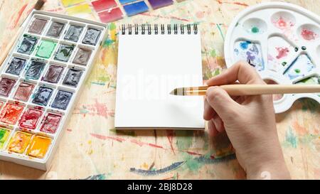 Künstler Malerei mit Aquarell Farbstoff Mockup flach legen. Draufsicht auf Maler Hand nehmen Aquarellfarbe in leerem Skizzenbuch. Stockfoto