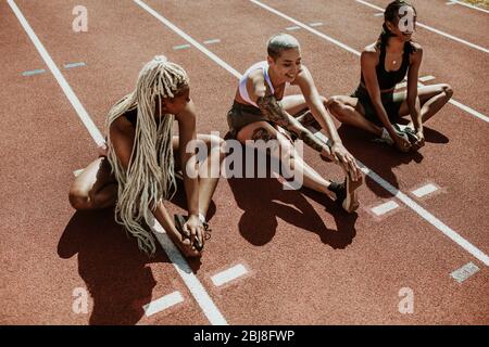 Drei Sportlerinnen sitzen auf einer Laufstrecke und strecken die Beine. Multiethnische Gruppe von Läufern, die im Stadion Aufwärmübungen machen. Stockfoto