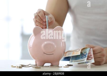Junger Mann, der Geld in ein Sparschwein auf einem Holztisch steckt. Stockfoto