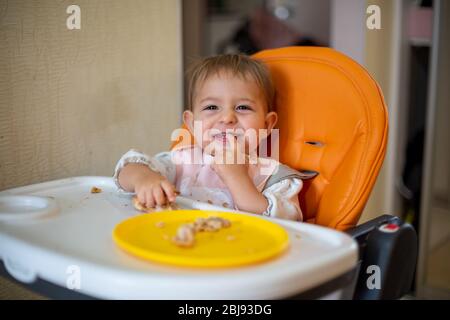 Niedliches Baby in einem orangefarbenen Kindersitz schaut die Kamera lachend an und zeigt Hand auf Zähne. Die Zähne sind gewachsen. Krümel und eine orangefarbene Platte mit einem Kuchen auf der t Stockfoto