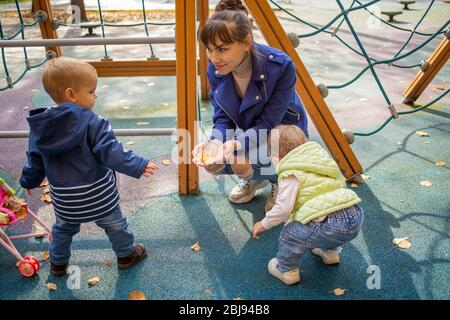 Eine junge schöne Mutter in einer blauen Jacke hockt in einem Herbstpark vor ihrem kleinen süßen Kleinkind Sohn und Tochter von 1 Jahr alt in blauer Kleidung Stockfoto