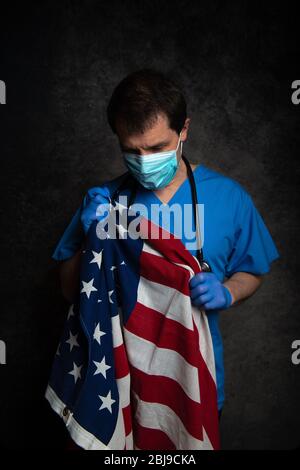 Traurig/nachdenklich, Arzt in blauem Krankenhauspeeling mit Gesichtsmaske und Stethoskop, Pflege der Stars & Stripes amerikanischen Flagge nahe seiner Brust. Stockfoto