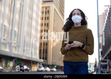 Kaukasische Frau, die eine Schutzmaske trägt und ihr Telefon auf der Straße benutzt Stockfoto
