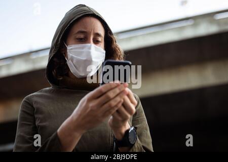 Kaukasische Frau, die eine Schutzmaske trägt und ihr Telefon auf der Straße benutzt Stockfoto