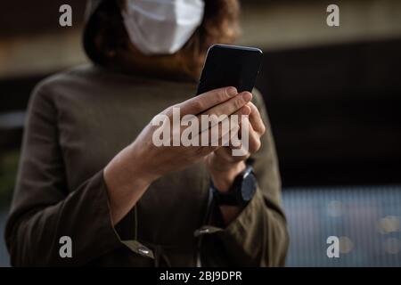 Kaukasische Frau, die eine Schutzmaske trägt und ihr Telefon auf der Straße benutzt Stockfoto