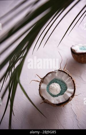 Zwei Hälften gebrochener Kokosnuss in ihren haarigen braunen Schalen und unfokussiertes grünes Palmblatt auf weiß strukturierter Oberfläche. Kokosnüsse sind mit blauer Kokosnuss gefüllt Stockfoto