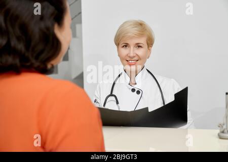 Selektiver Fokus der lächelnden Ärztin mit Stethoskop auf Hals mit Dokumenten und Blick auf die Kamera hinter der Rezeption in der Klinik. Freistellen Stockfoto