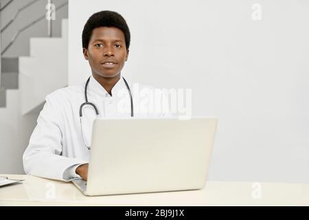 Porträt eines afrikanischen männlichen Arztes in weißem Laborkittel, der die Kamera anschaut. Professionelle Therapeutin mit Stethoskop am Hals posiert, mit Laptop an der Rezeption Stockfoto