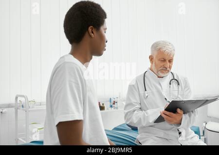 Schöner afrikanischer Mann trägt weißes T-Shirt auf Beratung mit Therapeut. Patient schaut auf den Arzt, wartet auf Ergebnisse. Eldery Arzt schreiben Presc Stockfoto