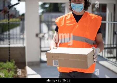 Junger Liefermann mit Paketbox im Außenbereich mit psa, Handschuhen und Gesichtsmaske, während der Sperrung der COVID-19 Pandemie. Fahrer mit Paket außen. Stockfoto