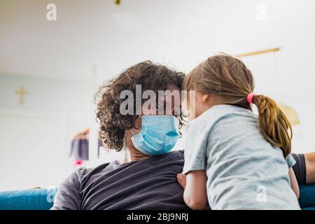 Kaukasischer Vater, trägt Gesichtsmaske, um Ansteckung zu vermeiden, scherzt mit Kind Mädchen während Covid-19 Pandemie Sperrung. Papa und Tochter schauen nah. Stockfoto