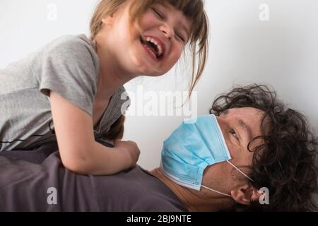Kaukasischer Vater trägt Gesichtsmaske, um Ansteckung mit lachenden Mädchen während Covid-19 Pandemie Sperrung zu vermeiden. Vater und Tochter Baby lachen und Stockfoto