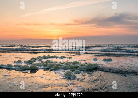 Sonnenuntergang über der Nordsee, von der niederländischen Küste aus gesehen. Meeresschaum wird an Land gewaschen. Stockfoto