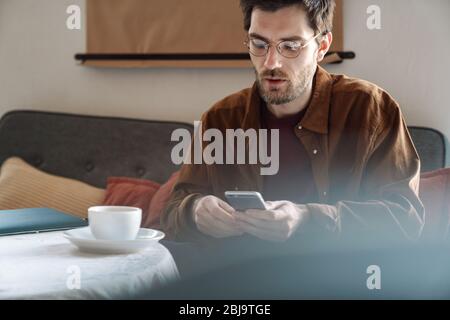 Bild von gutaussehenden fokussierten jungen Mann trägt Brillen mit Handy während der Ruhe in hellen modernen Café Stockfoto