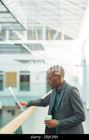 Afrikanischer junger Geschäftsmann, der Kaffee trinkt und ein Dokument liest, während er am Bürokorridor steht Stockfoto