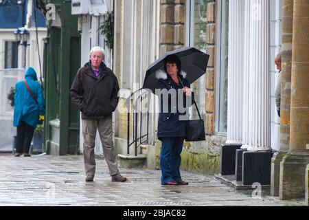 Bridport, Dorset, Großbritannien. April 2020. Wetter in Großbritannien. Shopper mit einem Regenschirm, der sich vor dem Regen in Bridport in Dorset schützt und an einem nassen Morgen vor einer Apotheke Schlange steht. Bild: Graham Hunt/Alamy Live News Stockfoto