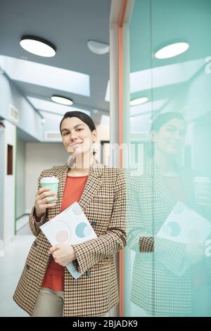 Porträt einer jungen Geschäftsfrau, die Dokument und eine Tasse Kaffee hält und die Kamera im Büro anschaut Stockfoto