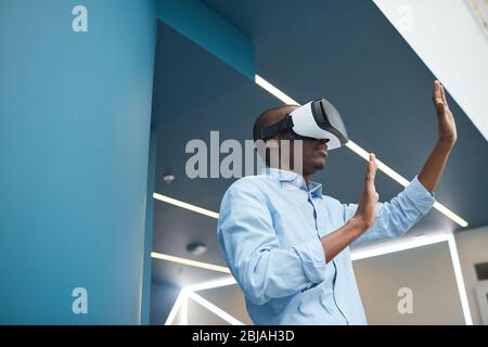 Afrikanischer junger Mann, der im Büro virtuelle Brille trägt und im Stehen und Gestik Stockfoto