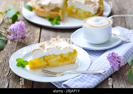 Köstliche Rhabarber-Kuchen mit süßem Baiser und einer Tasse Cappuccino auf einem Holztisch mit frühlingshafter lila Dekoration serviert Stockfoto