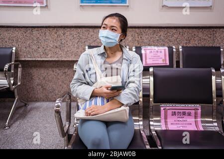 Junge asiatische Frau trägt chirurgische Gesichtsmaske sitzen in leeren Krankenhausbank mit Zeichen in Chinesisch Aufruf, soziale Distanz zu halten, in Hsichu, Taiwan Stockfoto