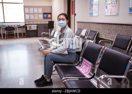 Junge asiatische Frau trägt chirurgische Gesichtsmaske sitzen in leeren Krankenhausbank mit Zeichen in Chinesisch Aufruf, soziale Distanz zu halten, in Hsichu, Taiwan Stockfoto