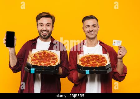 Zwei attraktive fröhlich aufgeregt jungen Männern Freunde in lässigen Kleidung stehen isoliert über gelben Hintergrund, zeigt leere Handy und Cred Stockfoto