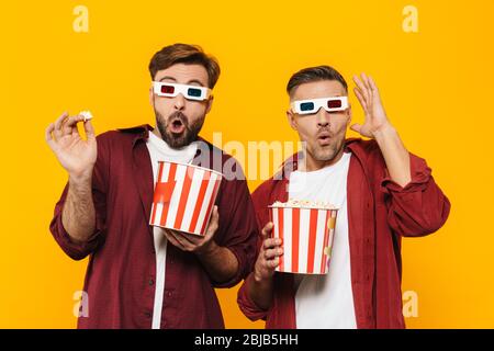 Zwei attraktive fröhlich aufgeregt jungen Männern Freunde tragen 3d-Brille, essen Popcorn stehend isoliert über gelbem Hintergrund Stockfoto