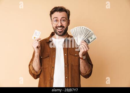 Bild eines fröhlichen, fröhlichen jungen, unrasierten Mannes, der auf einem beigefarbenen Wandhintergrund mit Geld und Kreditkarte isoliert ist. Stockfoto