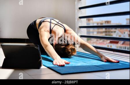 Frau Yoga Kind Pose auf Matte durchführen Online-Sitzung auf Tablet verwenden. Training zu Hause auf der Terrasse durch Selbstisolierung Pandemie Corona Virus Covid-19 Stockfoto