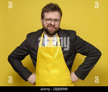 Seltsamer Mann in schwarzem Anzug und gelber Schürze steht und hält die Hände an den Hüften und starrt amüsant mit verengten Augen. Stockfoto