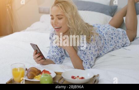 Fröhliche junge Frau, die Smartphone-Browsing und lachend auf dem Bett liegen Stockfoto