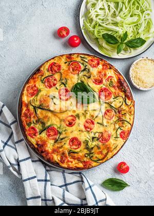 Zucchini-Kuchen und Zutaten auf grauem Hintergrund. Draufsicht auf köstliche herzhafte Kuchen mit Zucchini, Tomaten, Kräutern und Käse. Idee und Rezept für gesundes Backen und viel zu ernten von Zucchini. Vertikal Stockfoto