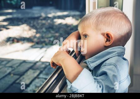 Kleiner Junge, der durch offenes Fenster nach draußen schaut. Bleiben Sie während der Quarantänesperre Corona COVID-19 zu Hause. Stockfoto