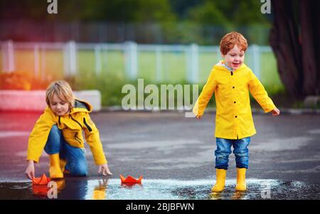 Nette Jungs, Brüder, die Spaß zusammen haben, die Papierboote in Frühlingspfützen starten, tragen Regenmäntel und Gummistiefel Stockfoto