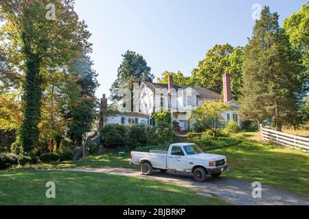 Ein Beat-up Pickup-Truck steht vor einem weißen Bauernhaus, umgeben von Bäumen und unter einem blauen Sommerhimmel, in Madison, CT, USA Stockfoto