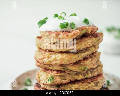 Zucchini-Fritten. Traditionelle Zucchini-Fritten in Stapel auf weißem Hintergrund. Vegetarische Zucchini-Pfannkuchen oder Fritten mit grünen Zwiebeln und Parmesan, serviert Sauerrahm oder griechischem Joghurt Stockfoto