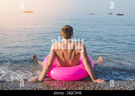 Glücklicher Mann mit großen rosa aufblasbaren Ring am Strand im Sommer sonnigen Tag. Rückansicht. Stockfoto