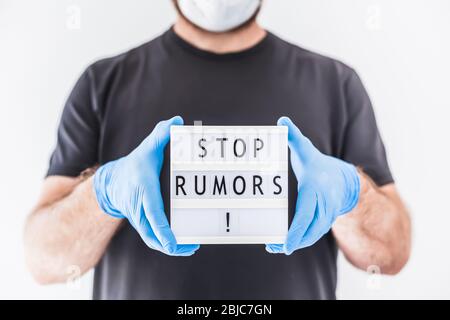 Fake News Infosemics during Covid-19 Pandemie Concept. Mann trägt Schutzmaske und medizinische Handschuhe an den Händen halten Leuchtkasten mit Text Stop Rumors. Stockfoto