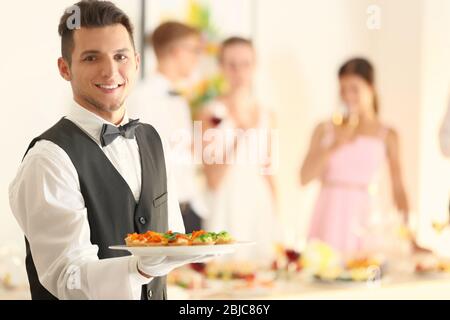 Schöner junger Kellner mit Teller voller leckerer Törtchen auf der Party Stockfoto