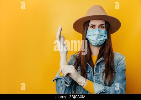 Attraktive junge Frau in Hut trägt schützende Gesichtsmaske, zieht medizinische Handschuhe an, Vorbereitung für eine gefährliche Reise während des Ausbruchs eines Coronavirus Stockfoto