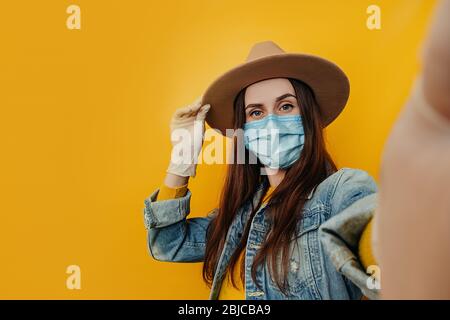 Junge attraktive weibliche Reisende in Hut und Handschuhe macht Selfie vor gelbem Hintergrund, in Denim Jacke gekleidet, trägt schützende medizinische Maske zu p Stockfoto