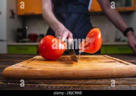 Koch schneidet eine rote Tomate in zwei Hälften mit einem großen Messer in Bewegung. Wasser- und Saftspritzer fliegen in verschiedene Richtungen und frieren in der Luft ein Stockfoto